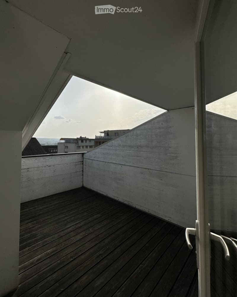 Modern wooden balcony with white concrete walls and a glass door leading inside, offering a view of the cityscape.