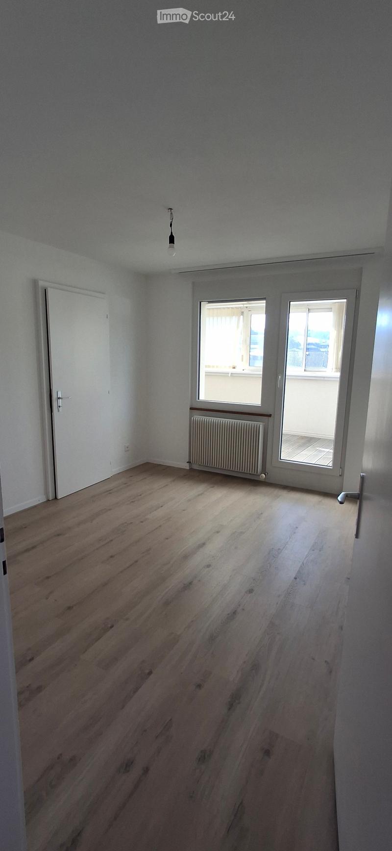 Empty room with wooden flooring, white walls, a radiator, and a door leading to a balcony area with windows.