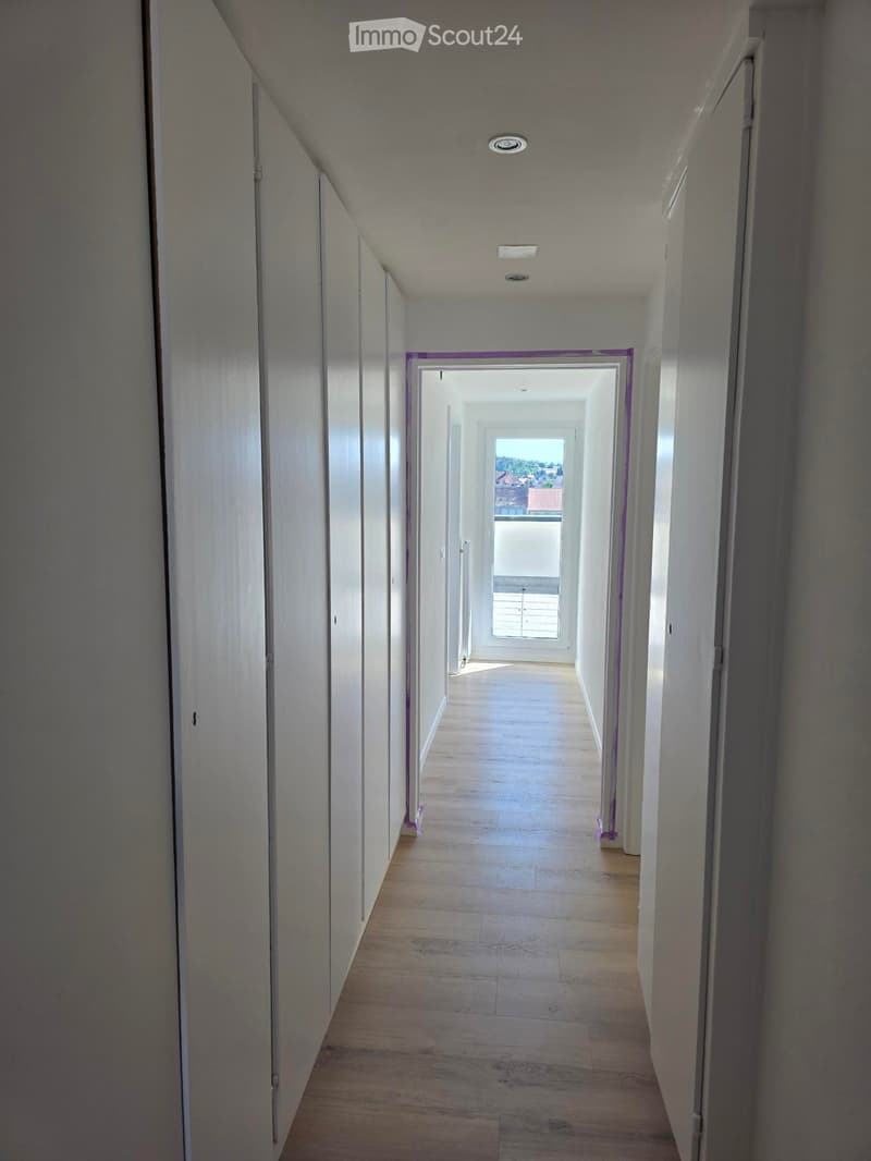 Empty hallway with wooden flooring, white walls, and recessed lighting. Doors line both sides, leading to an open area with a view of the outdoors.