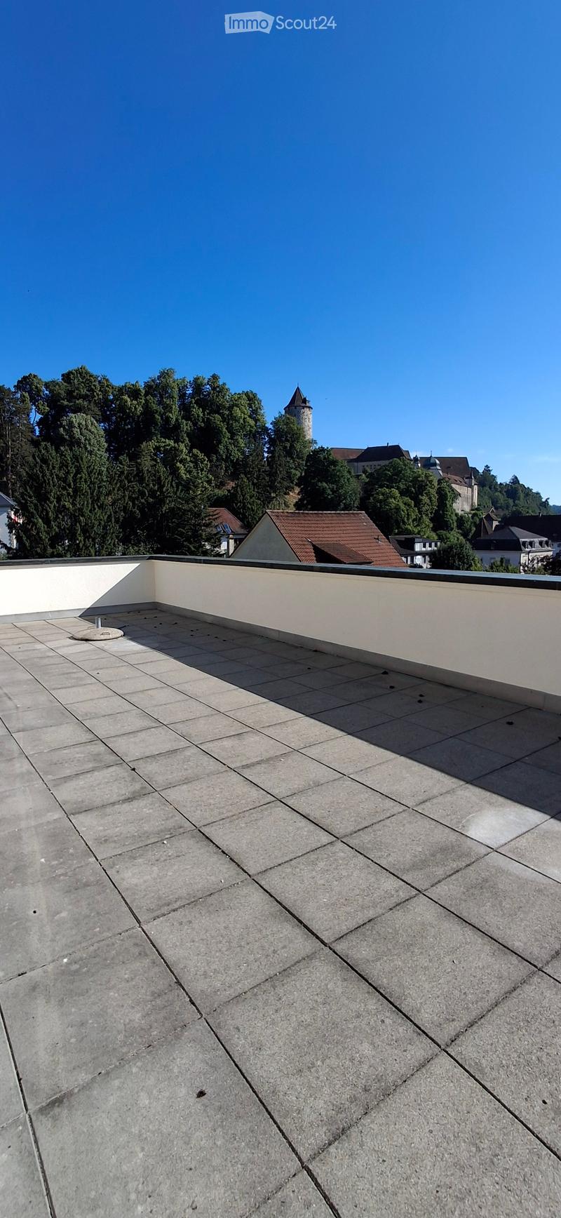 Flat rooftop terrace with concrete tiles, low white walls, and a view of surrounding buildings and greenery.