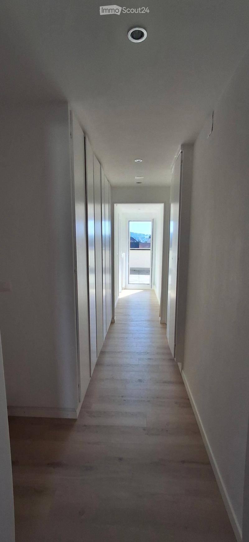 Empty hallway with light-colored walls, wooden flooring, and recessed ceiling lights. The hallway leads to an open doorway with a view of a balcony or outdoor area.