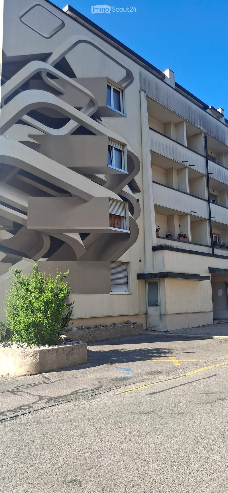 Modern multi-story building with curved balconies and large windows, featuring a small garden area in front.