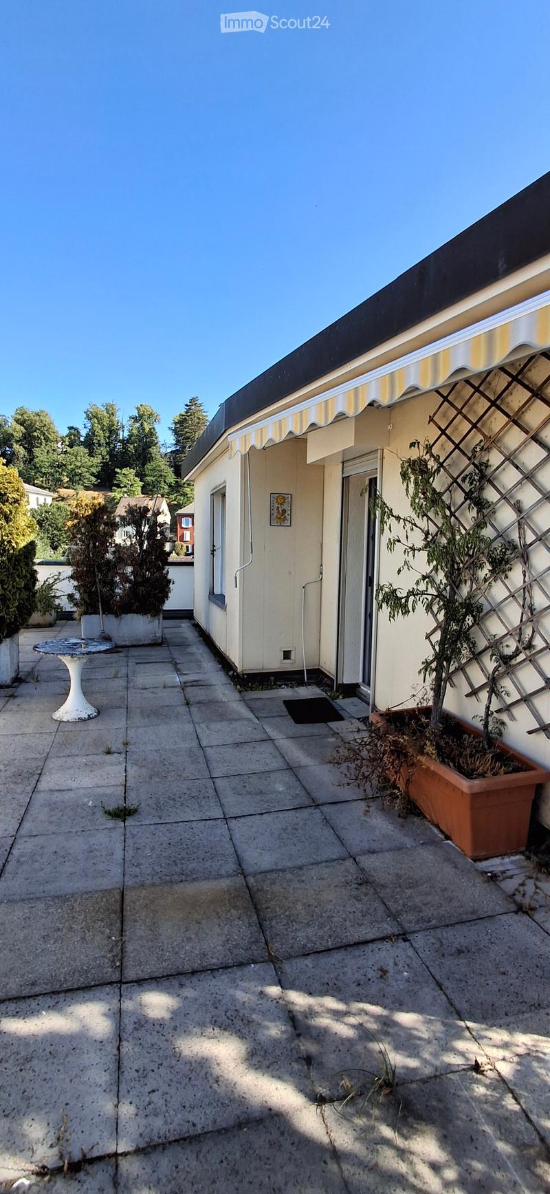 Concrete patio with a small table, potted plants, and a building entrance with a striped awning.