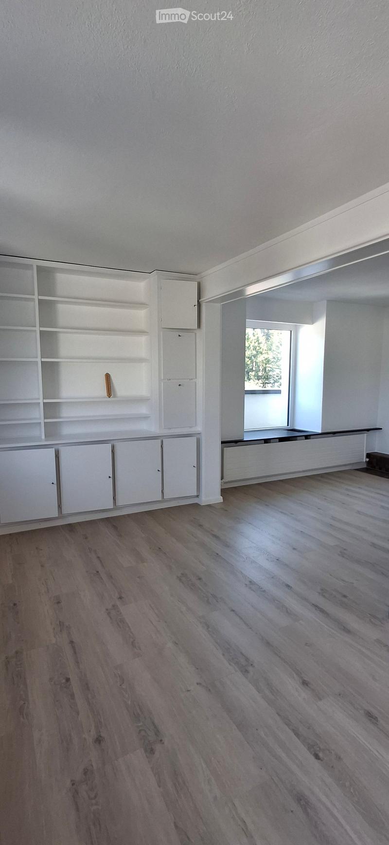 Empty room with light wooden flooring, white walls, built-in white shelving unit, and a large window letting in natural light.