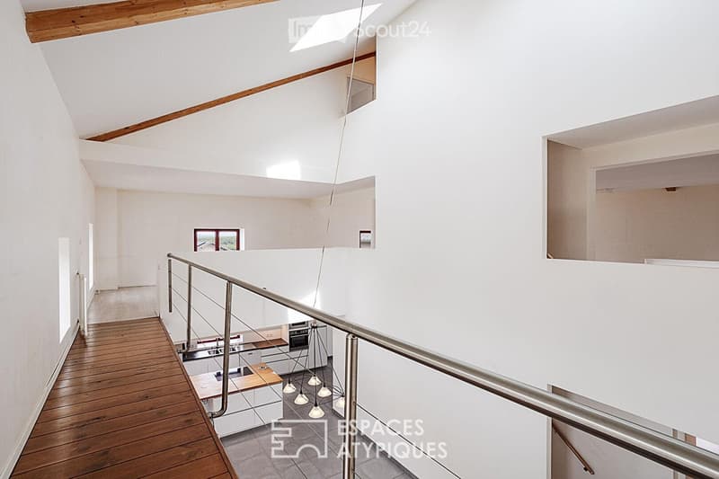 Open, modern corridor with wooden flooring, white walls, and a metal railing leading to a kitchen area below. The space features large windows and a skylight, providing ample natural light.
