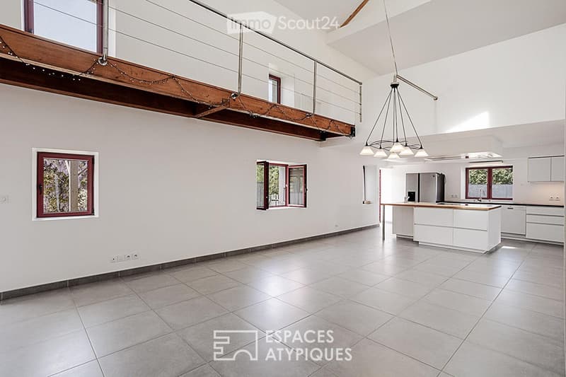 Bright, open-plan living and dining area with tiled flooring, white walls, and large windows. A modern kitchen with white cabinets and a central island is visible, along with a refrigerator and sink.
