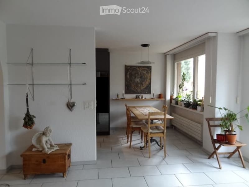 Bright, minimalist dining area with a wooden dining table and chairs, a small wooden stool with a decorative sculpture, potted plants on a side table, and a wall-mounted art piece.