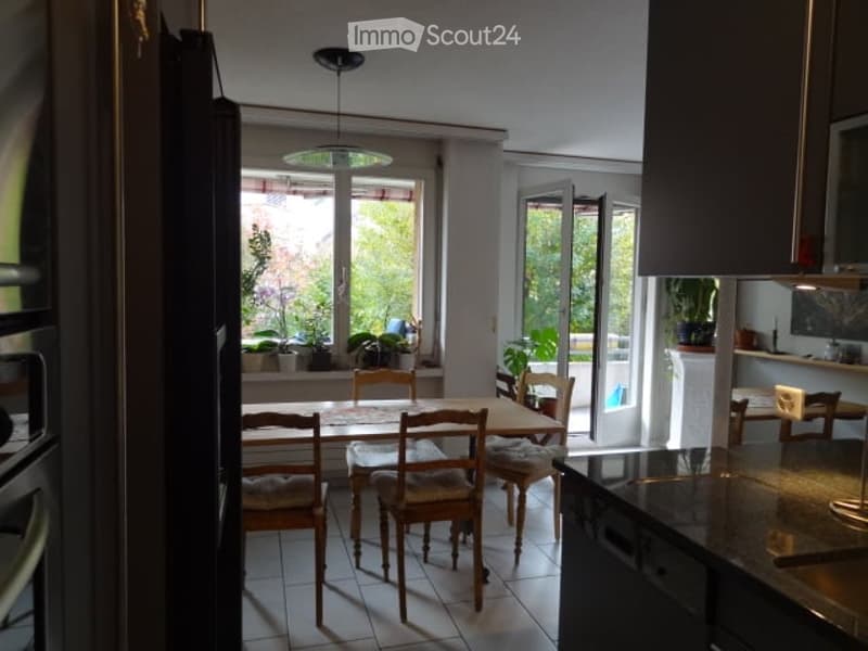 Bright, open-plan dining and kitchen area with large windows offering a view of greenery. The space features a wooden dining table with six chairs, modern kitchen cabinets, and a refrigerator. Potted plants are placed near the windows, adding a touch of n