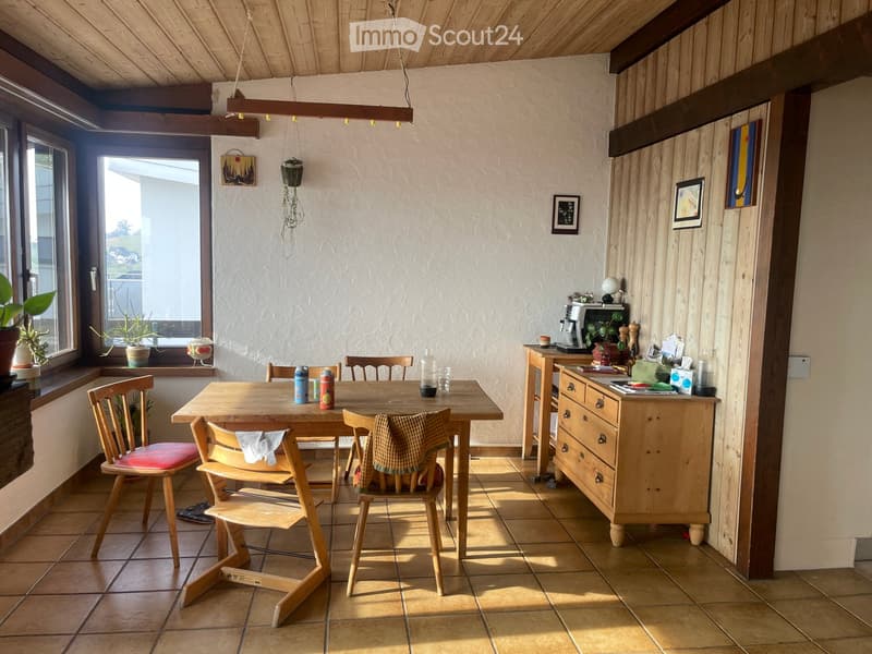 Bright kitchen with wooden table and chairs, tiled floor, and wooden cabinetry. Features a coffee maker, fridge, and various kitchen items on the counter.