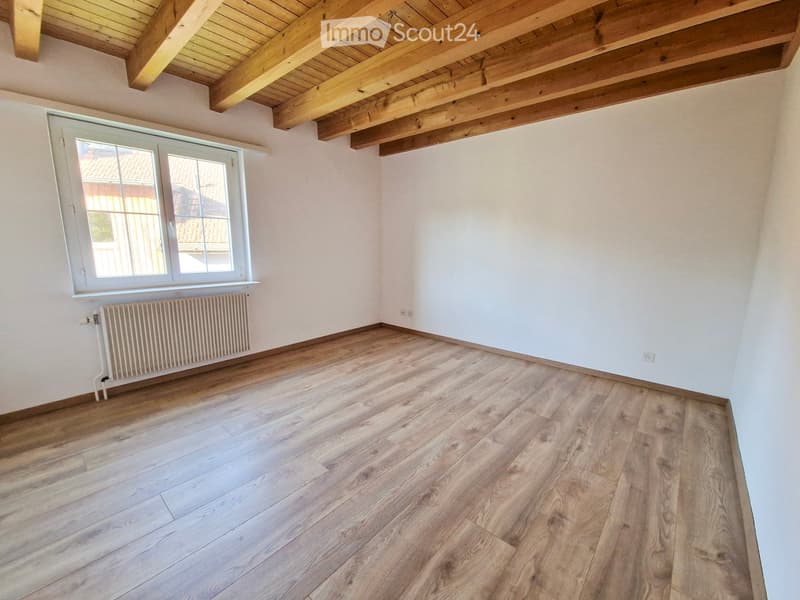 Empty room with wooden flooring, white walls, exposed wooden ceiling beams, and a single window with white frame. Radiator is visible under the window.