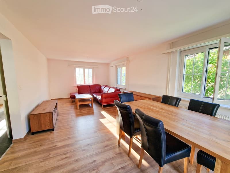 Bright living and dining area with wooden flooring, red leather sofa, coffee table, dining table with chairs, and large windows letting in natural light.