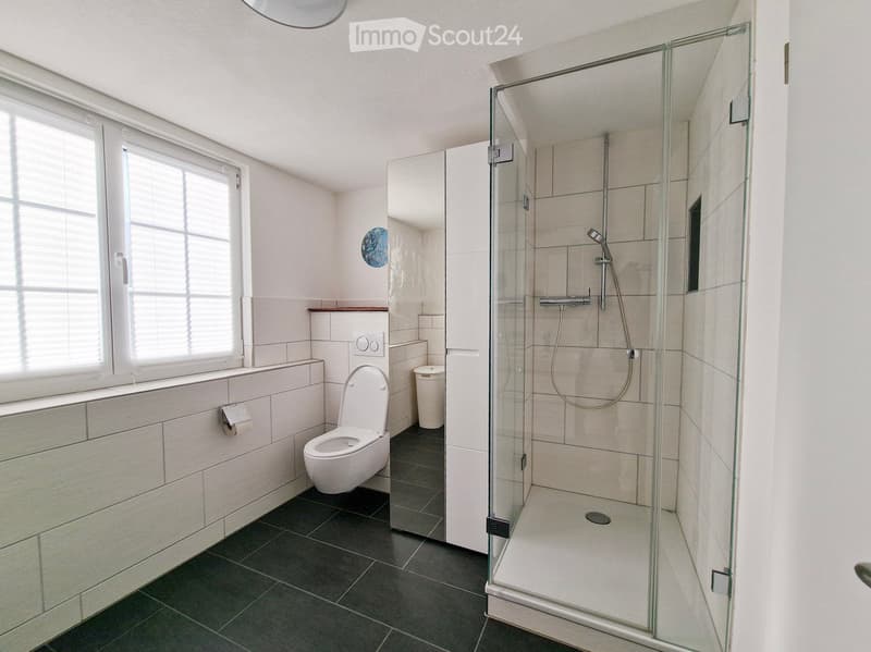 Modern bathroom with a toilet, glass-enclosed shower, and a window with blinds. The walls are tiled, and the floor is dark grey.