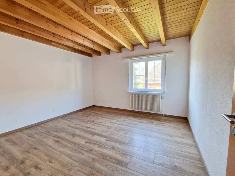Empty room with wooden flooring, white walls, exposed wooden ceiling beams, and a window with white frame and radiator below.