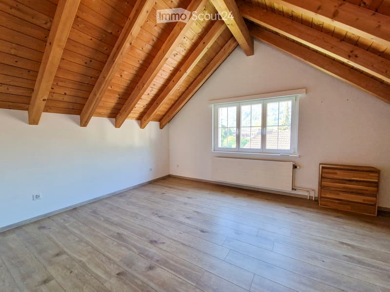 Empty room with wooden slanted ceiling, wooden floor, white walls, and a window with white frame. Radiator is visible under the window.