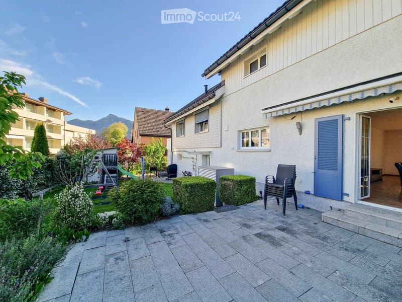 A well-maintained garden area with a patio, neatly trimmed hedges, a swing set, and outdoor furniture.