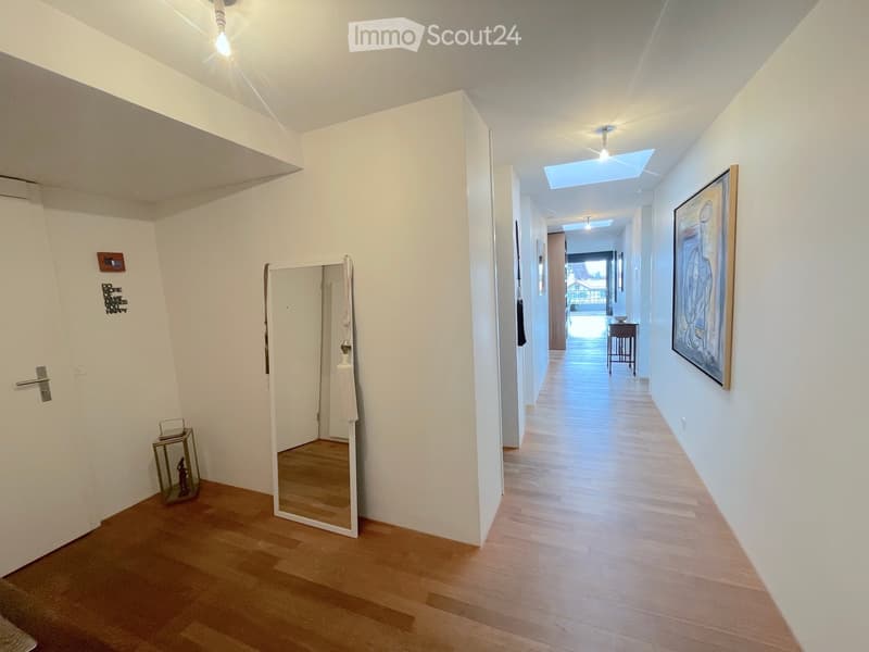 Bright hallway with wooden flooring, white walls, and modern lighting fixtures. Features a mirror, small decorative table, and artwork on the walls.
