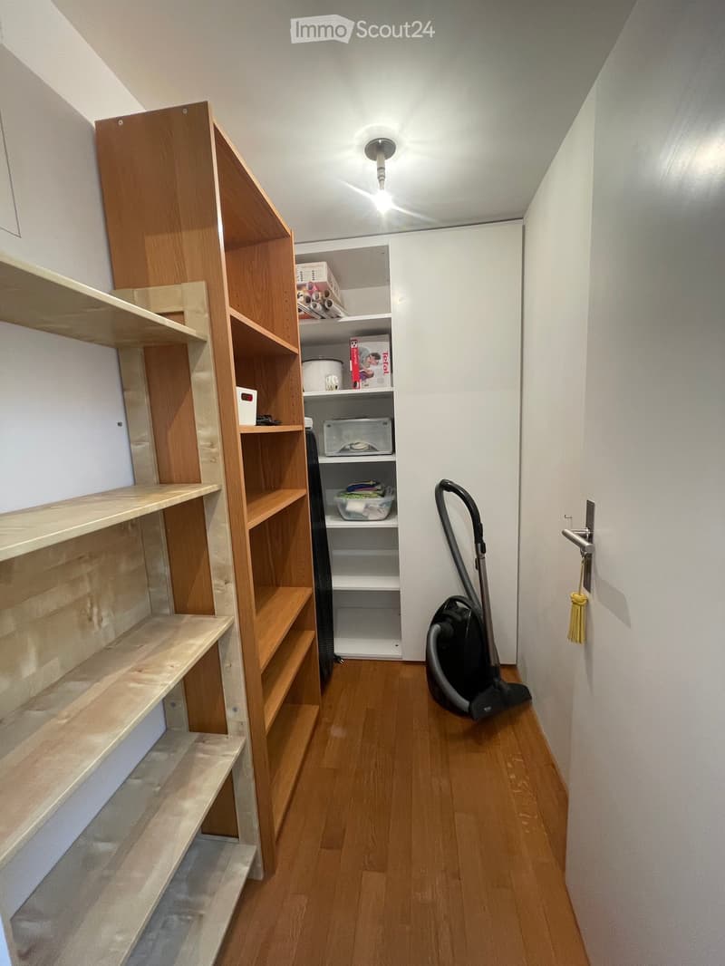 A narrow hallway with wooden flooring, white walls, and a built-in wooden shelving unit on the left side. A vacuum cleaner is placed on the floor near the door on the right.