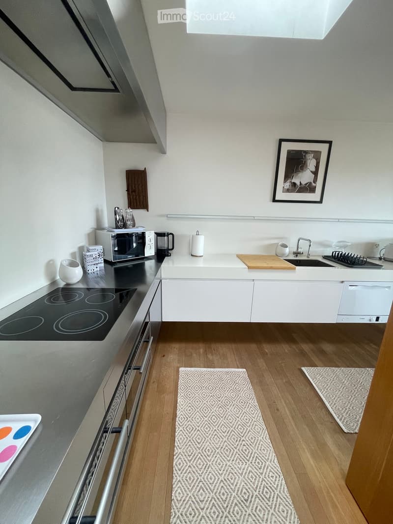 Modern kitchen with white cabinets, wooden flooring, induction stove, microwave, coffee maker, sink, and various small appliances. Features a framed picture on the wall and two patterned rugs on the floor.