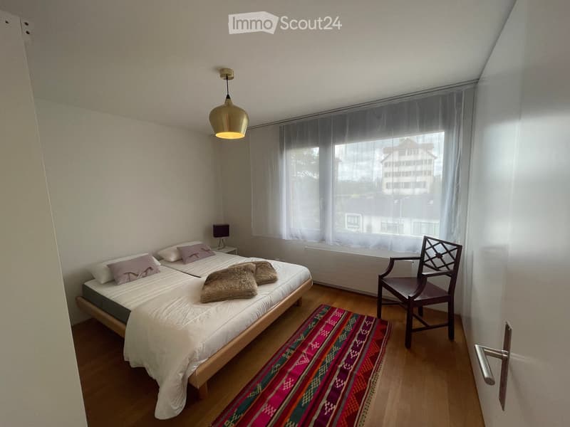 Cozy bedroom with a double bed, two pillows, a chair, a small table lamp, and a colorful rug on the floor.