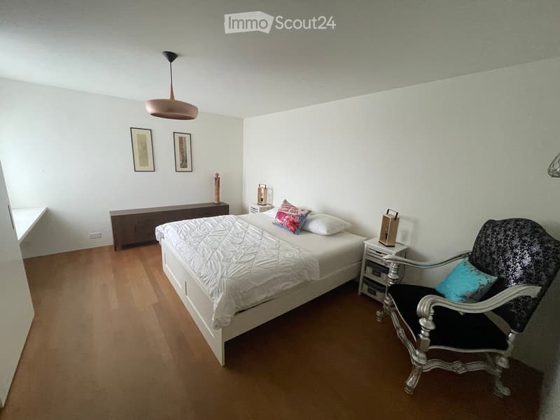A modern bedroom with a white bed, a black and white patterned chair, a nightstand with a lamp, and a small table with a stool. The room features wooden flooring and white walls.