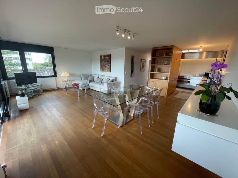 Open-plan living and dining area with wooden flooring, large windows, a white sofa, glass dining table with transparent chairs, a floor lamp, a TV stand, a bookshelf, and a kitchen area with cabinets and a vase with flowers on the counter.