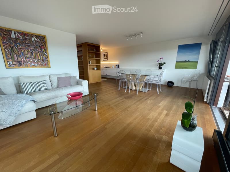 Modern living room with light wooden flooring, white sofa, glass coffee table, dining area with glass table and chairs, and decorative artwork on the walls.