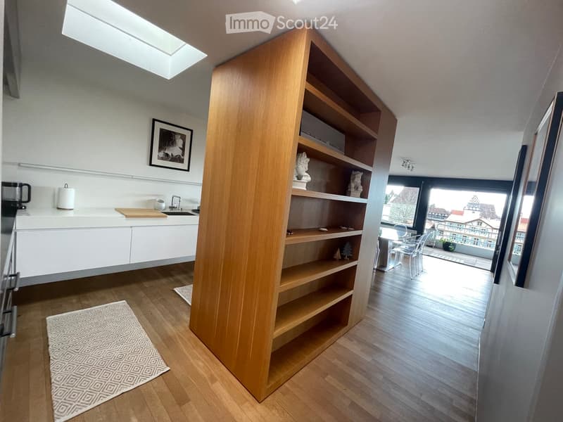 Modern kitchen with white cabinets, wooden shelves, and a large window overlooking a balcony. Includes a sink, cutting board, and decorative items on shelves.