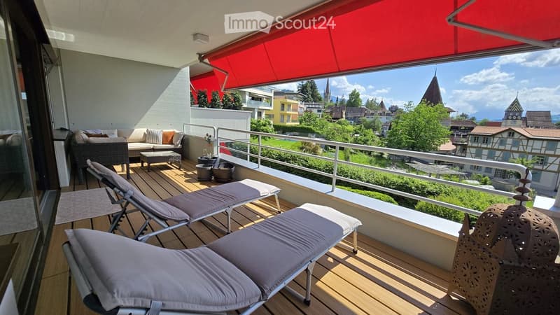Balcony with wooden decking, lounge chairs, sofa, coffee table, and potted plants, offering a scenic view of the town and surrounding landscape.