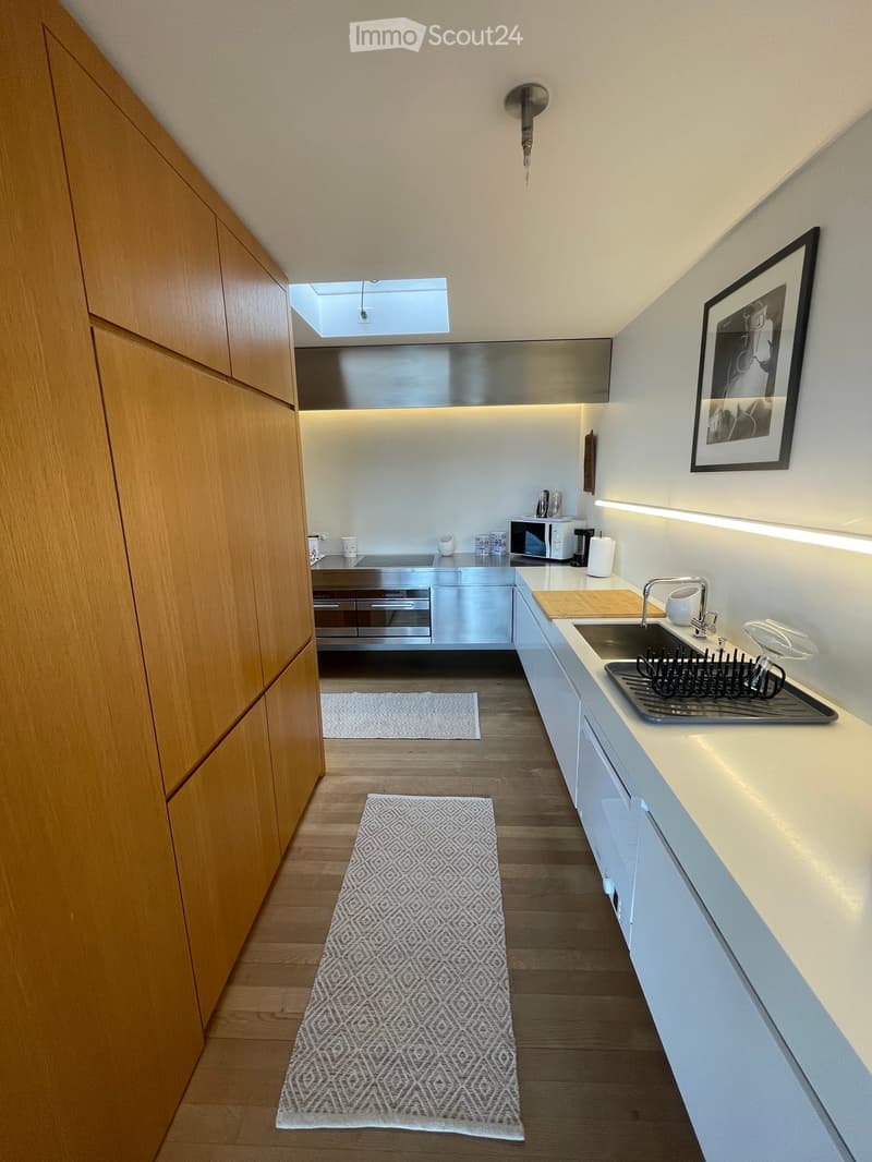 Modern kitchen with wooden cabinets, white countertops, stainless steel appliances, and a small sink area with a faucet and dish rack. The floor is wooden, and there are two small rugs placed on it.