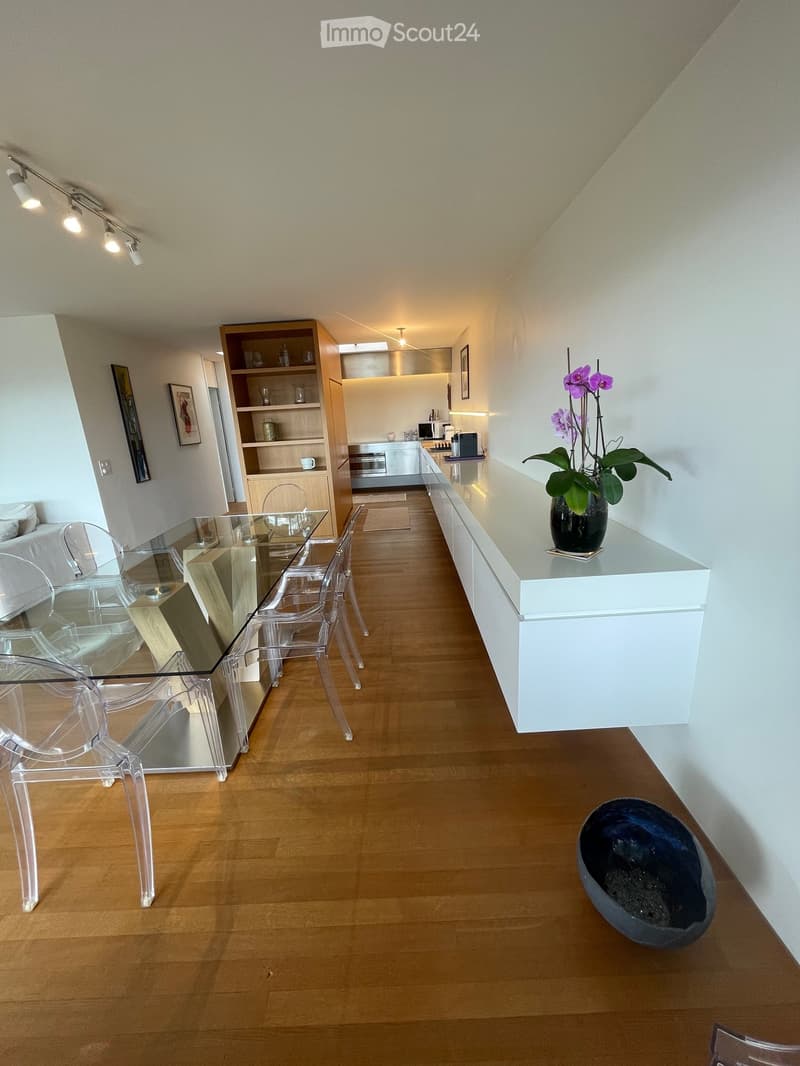 Modern open-plan living and dining area with a white sofa, glass dining table with transparent chairs, kitchen area with cabinets and appliances, and decorative elements including a flower vase and a bowl on the floor.