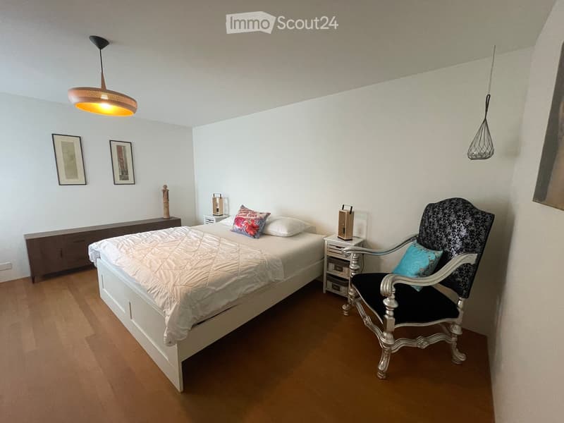A well-lit bedroom with a white bed, a decorative chair, nightstands, and artwork on the walls.