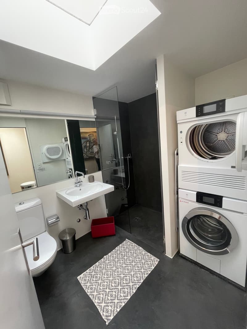 Modern bathroom with a toilet, sink, mirror, washing machine, and dryer. Features a dark grey floor and a patterned rug.