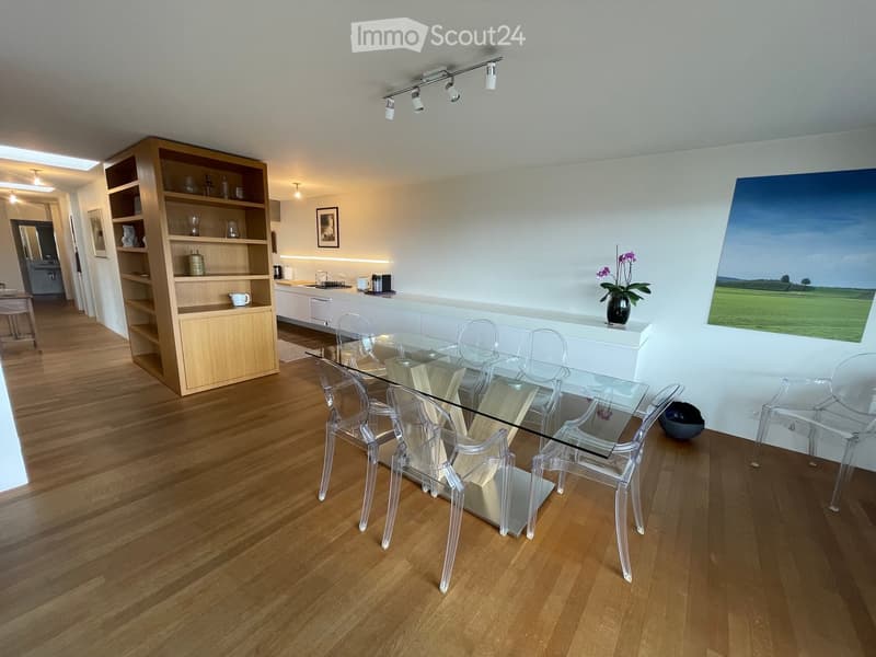Modern dining area with a glass table and transparent chairs, a kitchen counter, and a wooden cupboard with shelves.