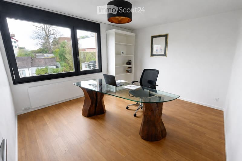 Modern office space with a glass desk, office chair, laptop, bookshelf, and a large window offering a view of the outdoors.