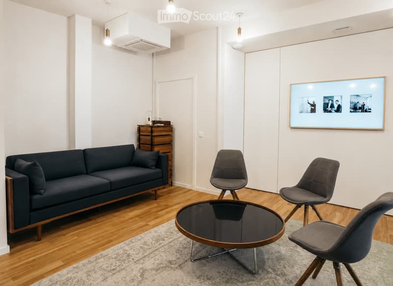 Modern living room with a dark blue sofa, glass coffee table, three grey chairs, a large screen displaying images, and a wooden cabinet.