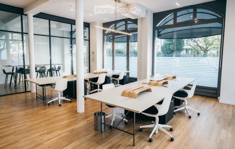 Modern office space with large windows, white tables, white chairs, and wooden stools arranged around tables.