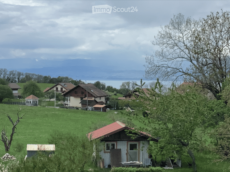 A rural landscape with scattered houses, green fields, and a distant lake under a partly cloudy sky.