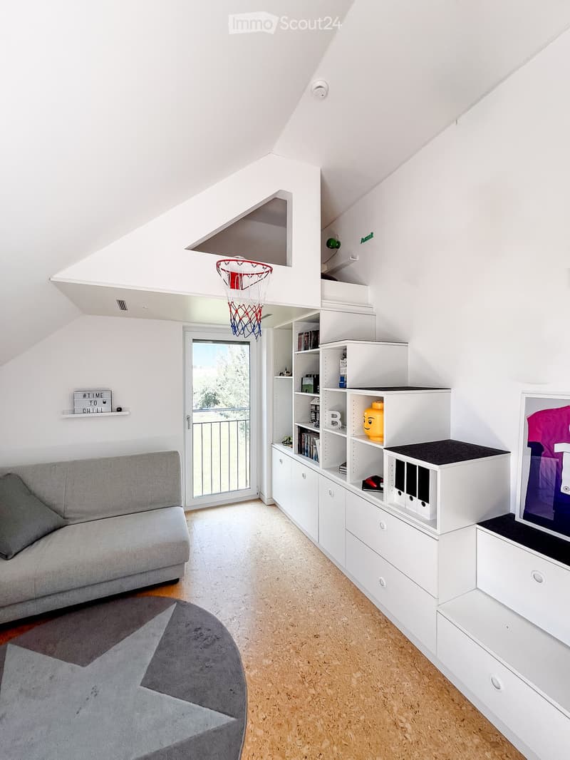 Modern attic room with slanted ceilings, white walls, and light wood flooring. Features a grey sofa, ottoman, built-in white shelving unit, and a small balcony with glass doors.