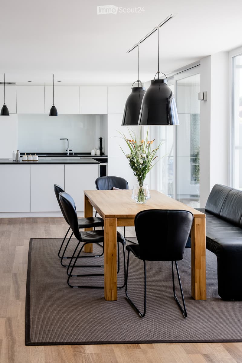 Modern open-plan living and dining area with a wooden dining table, black chairs, a black leather sofa, pendant lights, and a sleek kitchen area with white cabinets and a black countertop.