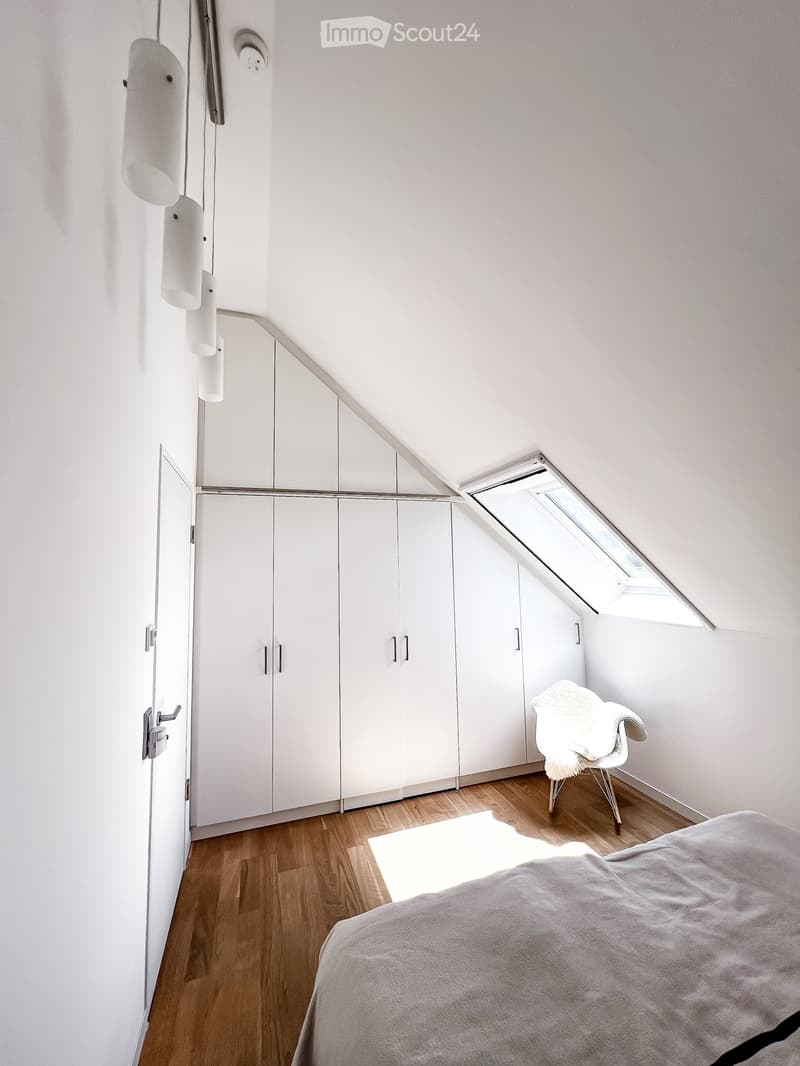 Modern bedroom with white walls, wooden floor, large built-in wardrobe, bed with grey duvet, white chair with fur throw, skylight window.