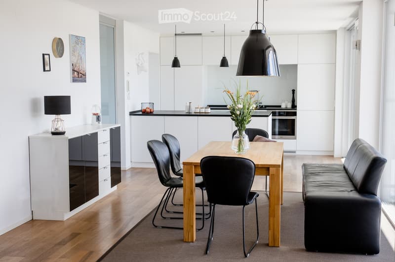 Modern open-plan living and dining area with a wooden dining table, black leather chairs, a black leather sofa, pendant lights, and a kitchen area with white cabinets and a black countertop.