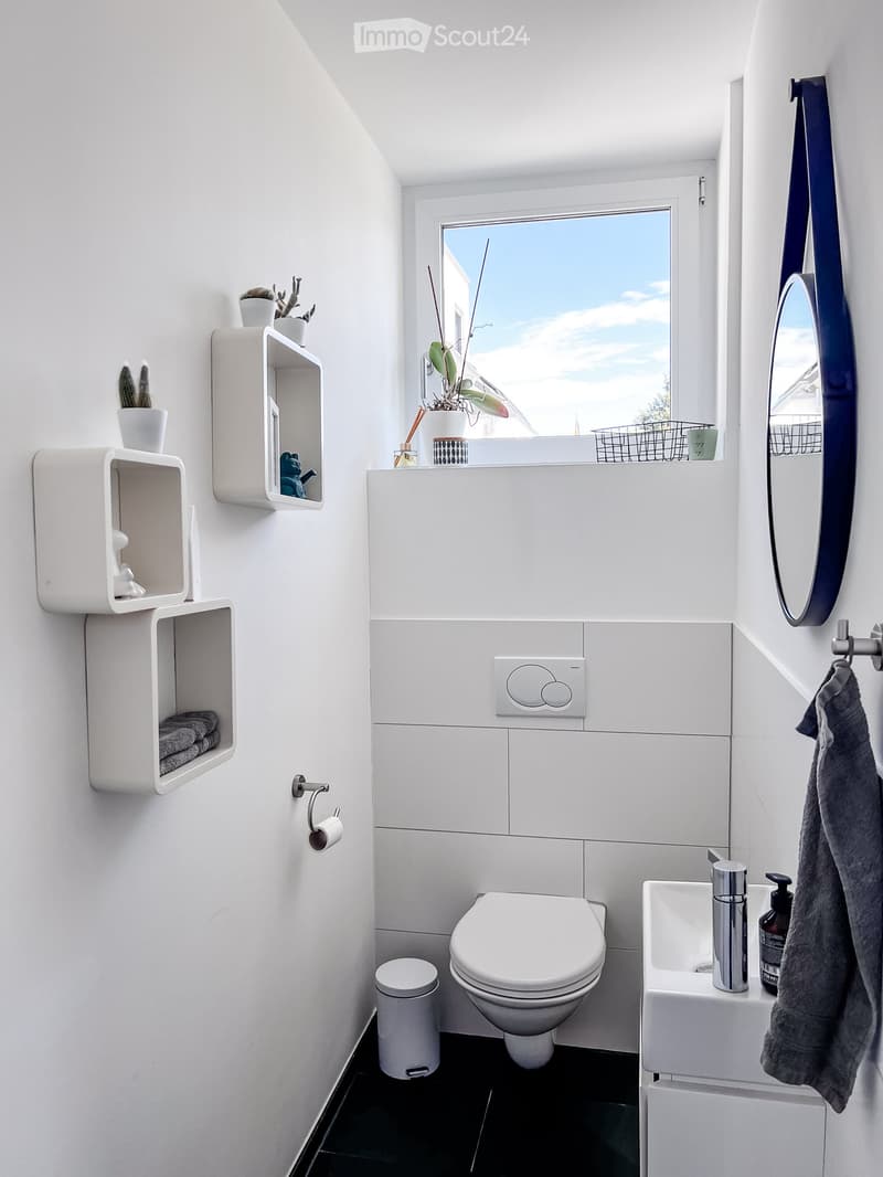 Modern bathroom with white tiles, a toilet, a sink, a mirror, wall-mounted shelves, and a window with a view of the sky.