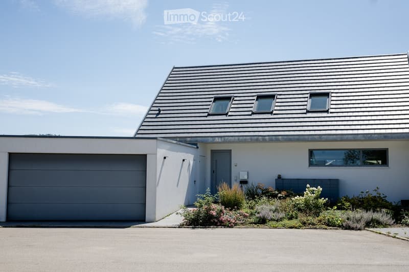 Modern two-story house with a grey garage, white walls, and a sloped roof with skylights. The front yard features a small garden with various plants and flowers.