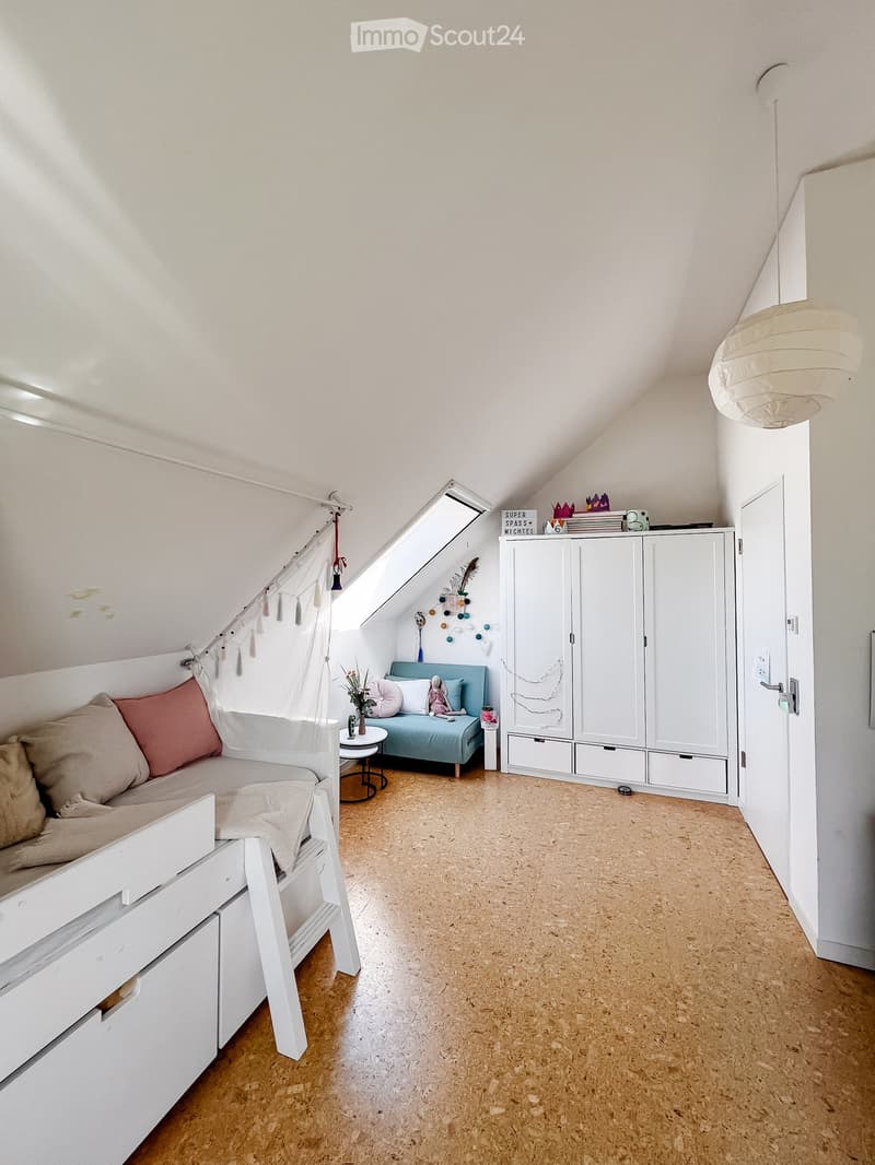 Cozy attic bedroom with a built-in bed, a small sofa, a round table, and a white cabinet. The room features wooden flooring and slanted ceilings with exposed beams.