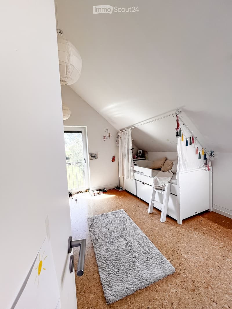 Cozy attic bedroom with slanted ceilings, white walls, wooden floor, grey rug, bed with white frame and pillows, nightstand, and a small window with a view of greenery.
