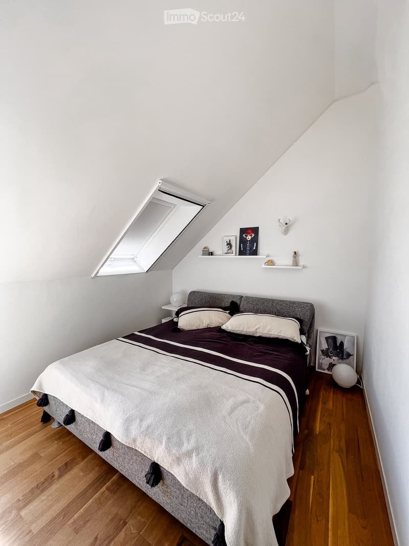 Bedroom with a grey headboard bed, dark brown and white bedding, wooden floor, shelves with decorative items, and a skylight window.