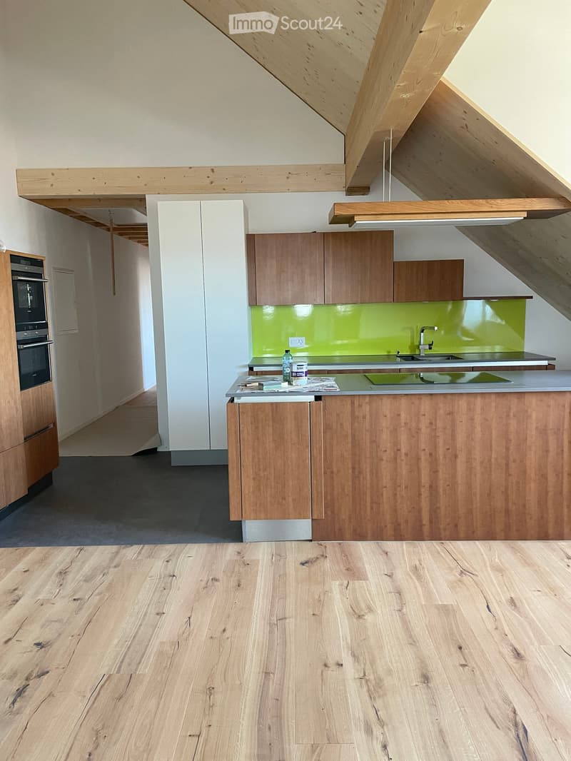 Modern kitchen with wooden cabinets, a central island, and a green backsplash. The floor is light wood, and the ceiling features exposed wooden beams.