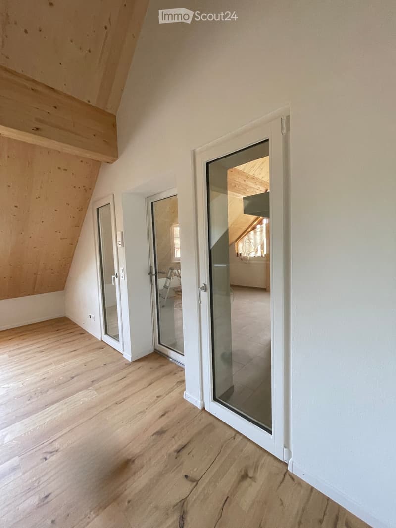 Empty attic room with wooden flooring, white walls, and large glass doors leading to another room.
