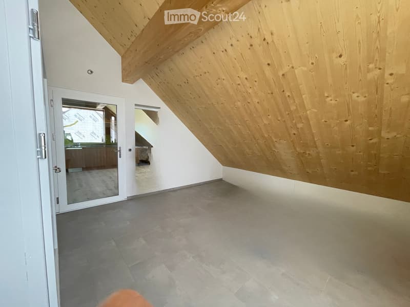 Empty room with wooden slanted ceiling, grey tiled floor, white walls, and a glass door leading to another room.