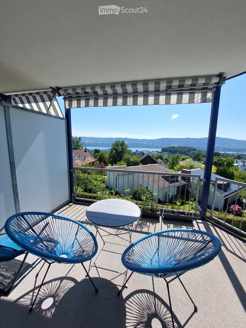 Balcony with a round table and two blue chairs, overlooking a scenic lake and mountain view.